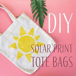 A white tote bag with a sun design printed on it over a pink background with green foliage.